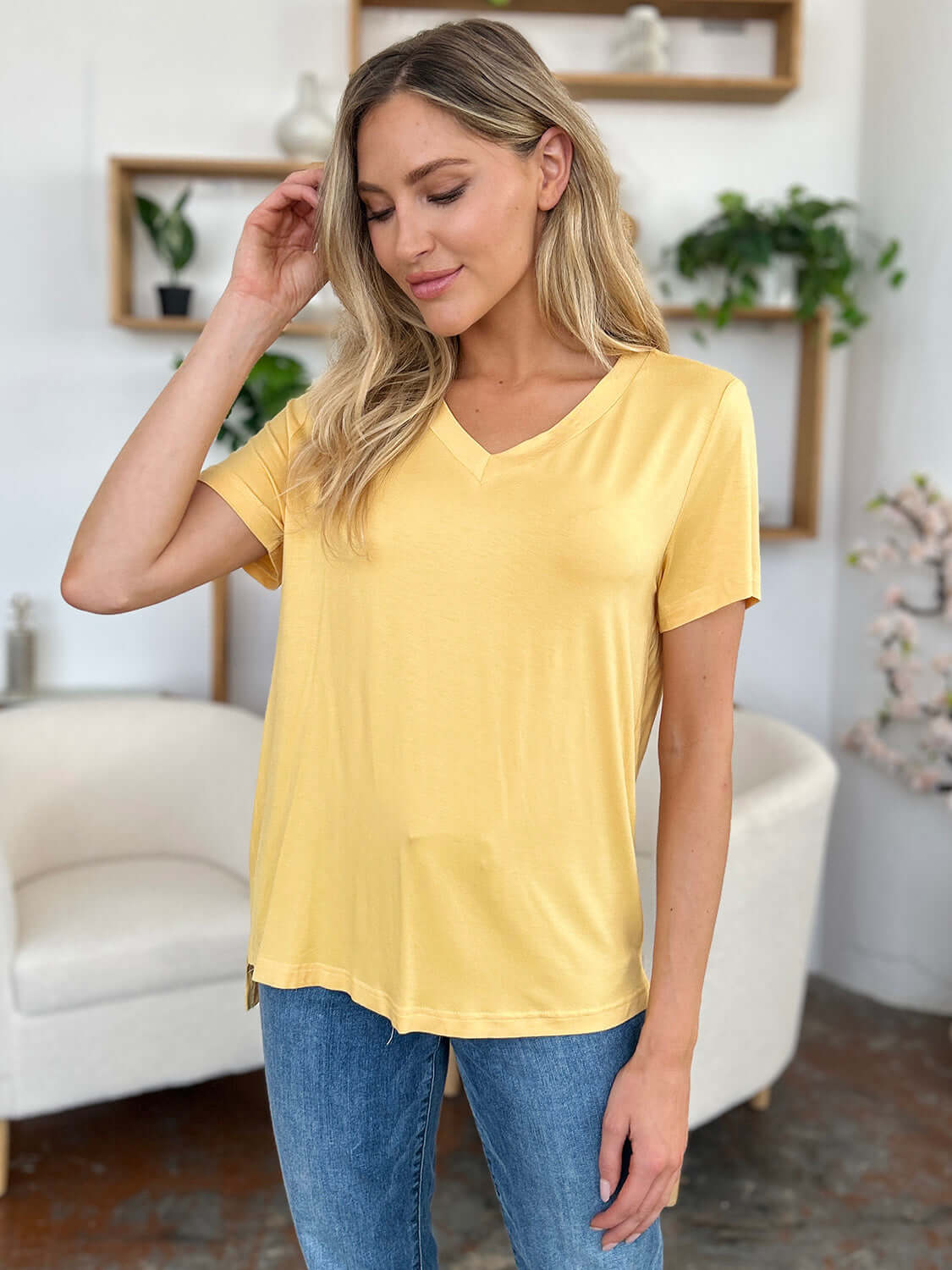 Woman wearing a bamboo V-neck high-low t-shirt in pale yellow, styled with blue jeans, standing in a cozy living room with plants and white furniture.