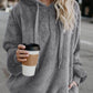 Stylish woman wearing a grey teddy hoodie with drawstring, holding a coffee cup in a city setting.