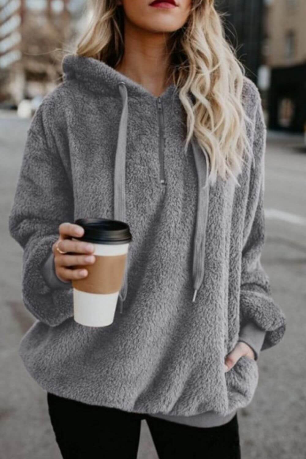 Stylish woman wearing a grey teddy hoodie with drawstring, holding a coffee cup in a city setting.