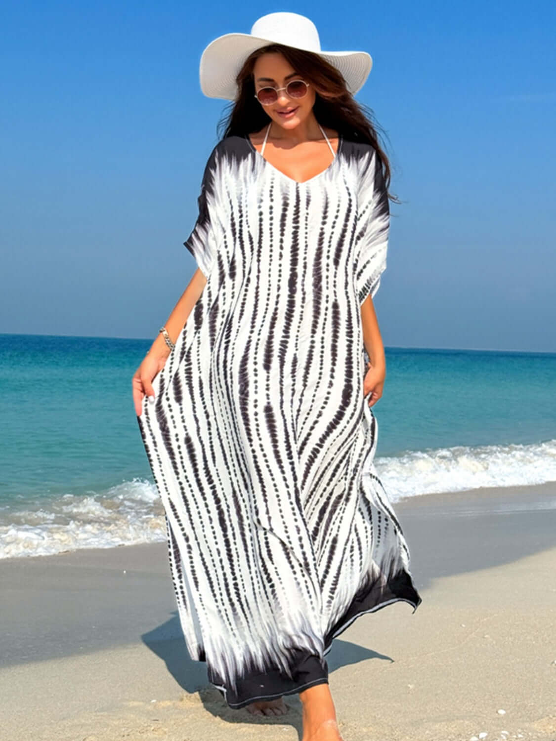 Woman in slit tie-dye half sleeve cover up on the beach, wearing a white hat and sunglasses, with ocean background.