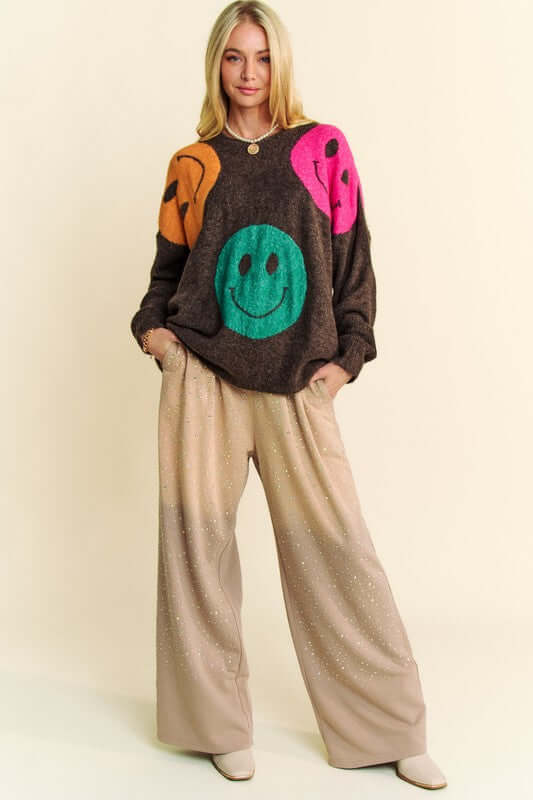 Trendy woman wearing Davi & Dani oversized sweater with colorful smiley faces, styled with relaxed pants for a cozy look.