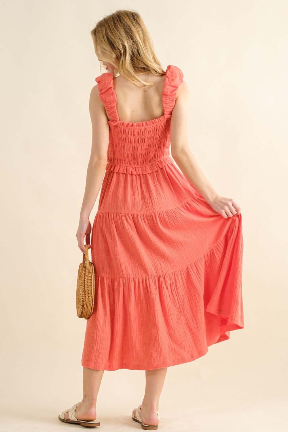 Woman wearing a coral smocked ruffled tiered dress holding a woven bag, showcasing the back and flowy silhouette of the dress.