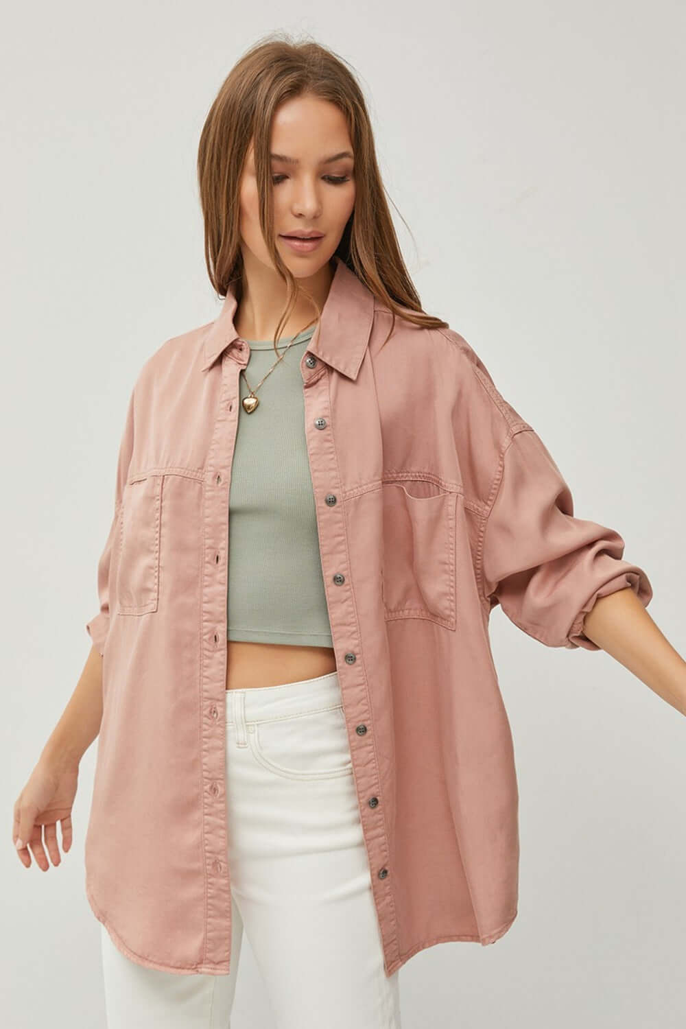 Woman wearing RISEN full-size button-up long sleeve denim shirt in pink, paired with a green crop top and white pants, casual stylish look