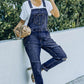 Woman wearing pocketed distressed denim overalls and sunglasses, leaning against a wall outdoors. Acid wash overalls with slight stretch.