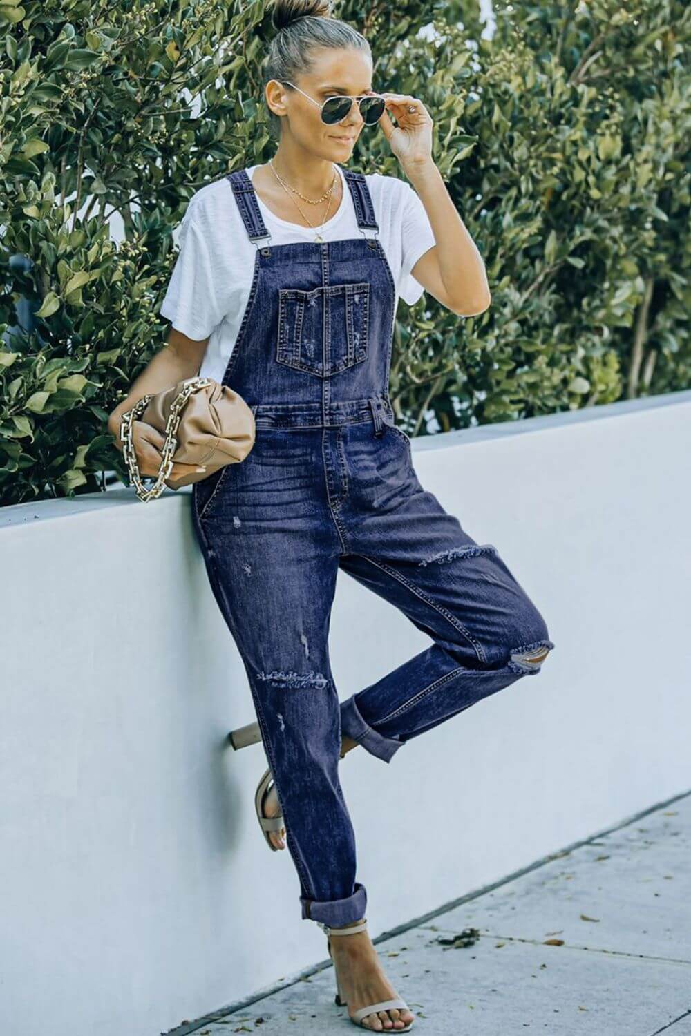 Woman wearing pocketed distressed denim overalls and sunglasses, leaning against a wall outdoors. Acid wash overalls with slight stretch.