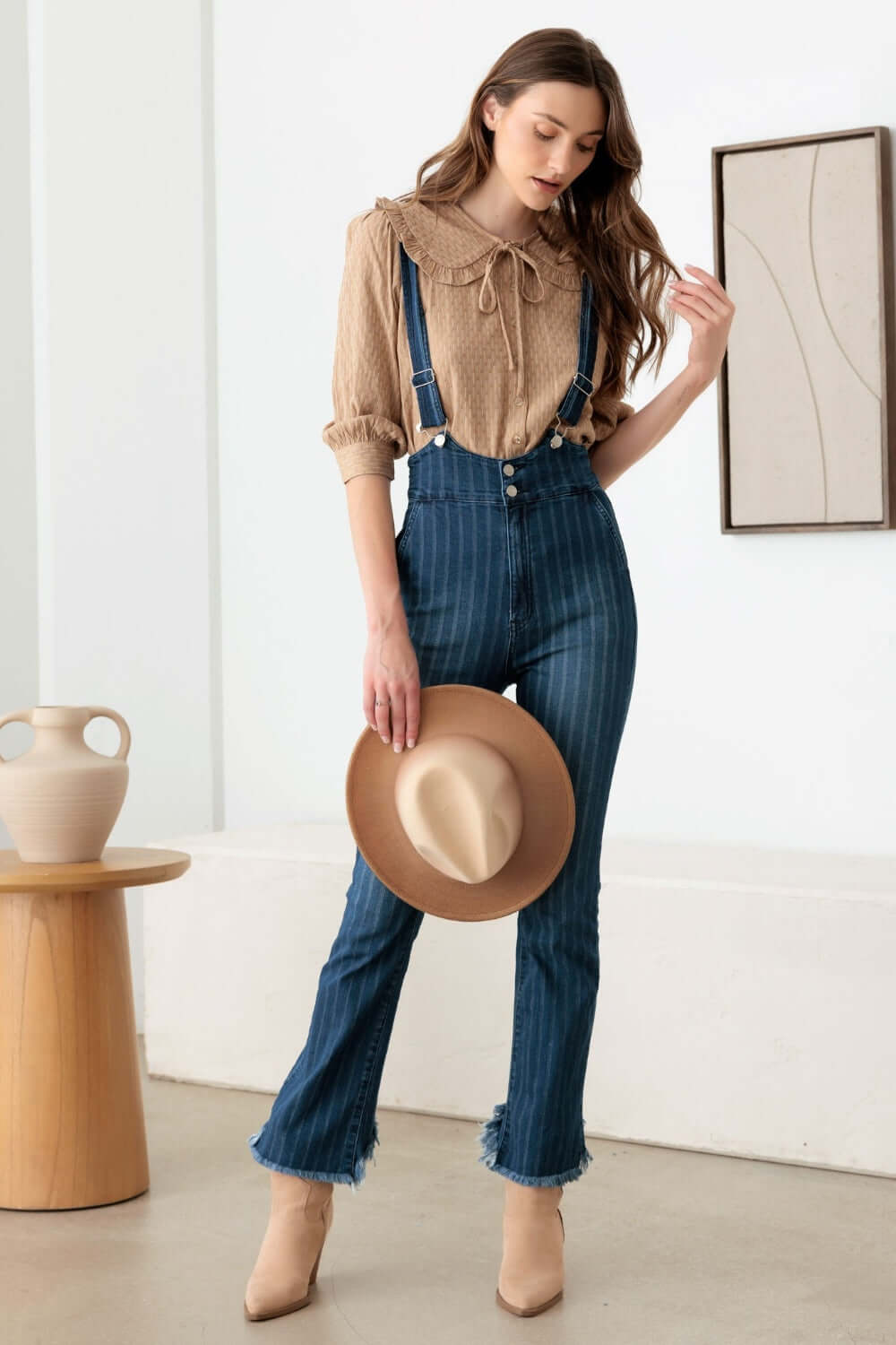 Woman wearing striped stretched suspender denim overalls with high waist, front pockets, zip closure, and flare fit, holding a hat.