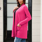 Woman wearing a pink open front long sleeve cardigan with pockets, styled with jeans, standing in front of a black door and window.