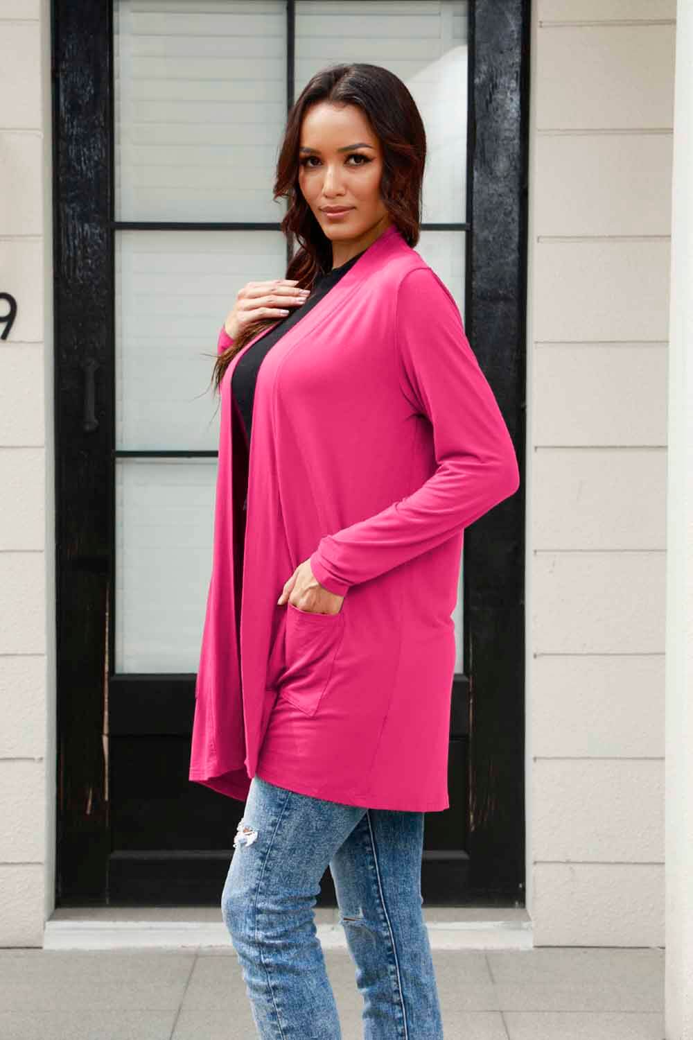 Woman wearing a pink open front long sleeve cardigan with pockets, styled with jeans, standing in front of a black door and window.