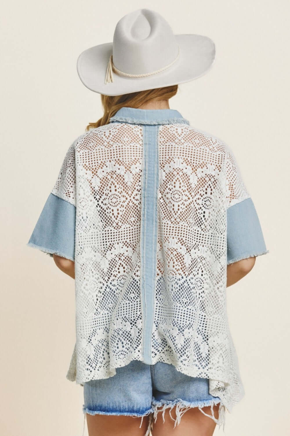 Back view of woman wearing a button-up short sleeve lace shirt with denim accents and a white hat, paired with denim shorts.