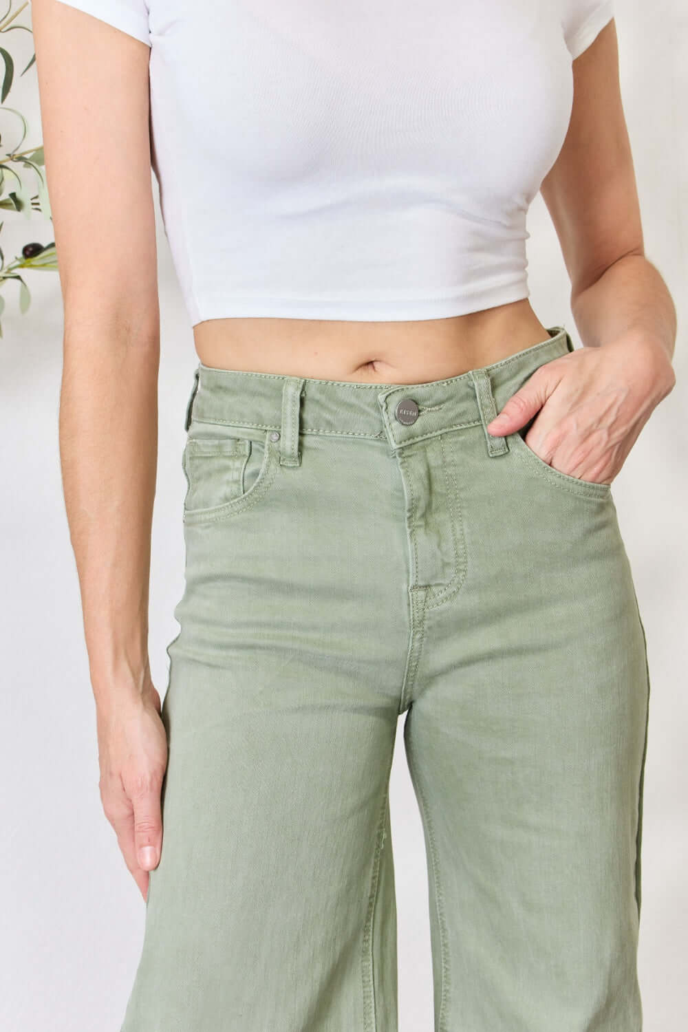 Woman wearing high-waisted raw hem wide-leg jeans in a soft green color, paired with a white crop top. Risen Jeans for a trendy look.