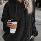 Woman in a cozy black teddy hoodie holding a coffee cup on a city street, showcasing style and comfort.