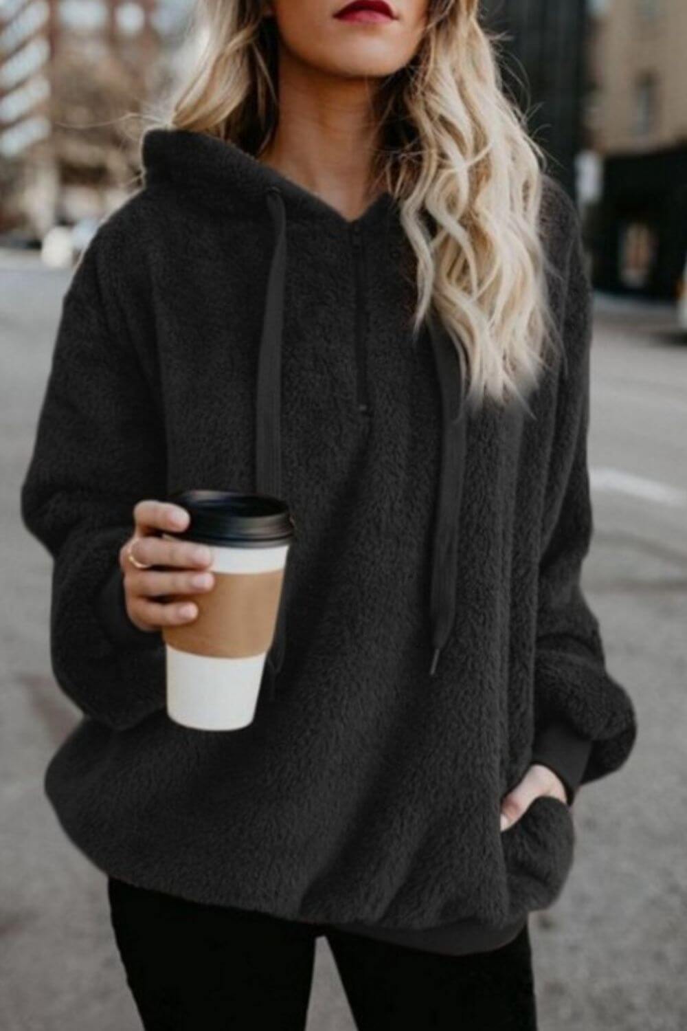 Woman in a cozy black teddy hoodie holding a coffee cup on a city street, showcasing style and comfort.