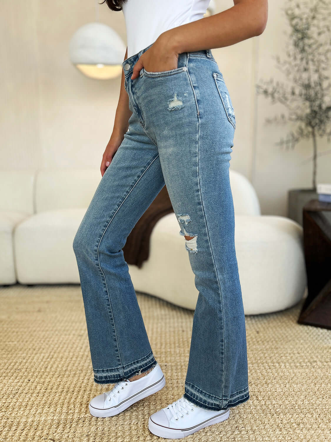 Woman wearing Mid Rise Destroyed Hem Distressed Jeans by Judy Blue, paired with a white tee and sneakers in a cozy living room.