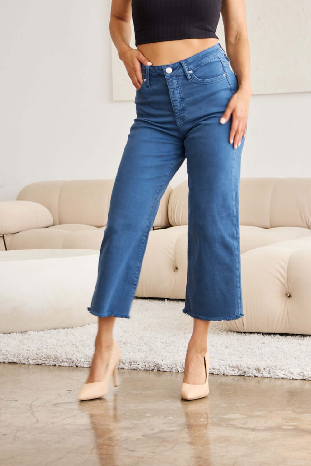 Woman wearing Crop Chloe Full Size Tummy Control High Waist Raw Hem Jeans by RFM Jeans, paired with beige heels in a stylish living room setting.