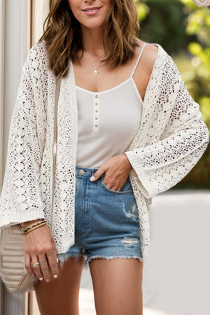 Woman wearing openwork open front dropped shoulder cardigan over white tank top and denim shorts.