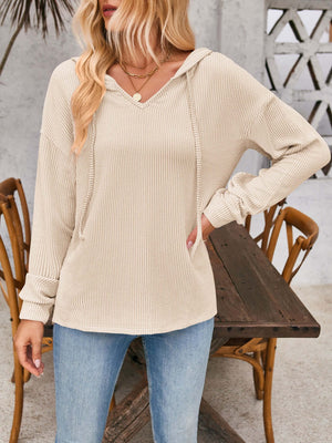 Woman wearing a beige drawstring dropped shoulder long sleeve hoodie with denim jeans, standing by wooden chairs and table outdoors.