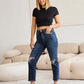 Woman wearing Crop Dylan Full Size Tummy Control Distressed High Waist Raw Hem Jeans by RFM Jeans, standing in a stylish living room.