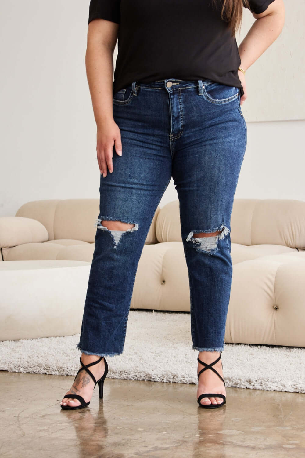 Woman wearing RFM Jeans distressed high waist raw hem jeans with tummy control, pairing with black top and strappy heels.