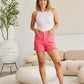 Woman wearing pink high waist raw hem denim shorts and a white tank top standing in a stylish living room.