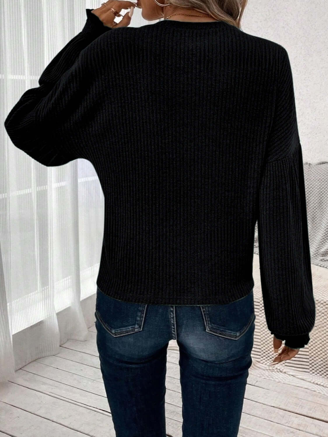 Rear view of woman wearing Bella Road Ribbed Round Neck Lantern Sleeve T-Shirt in black with jeans, standing by a window.