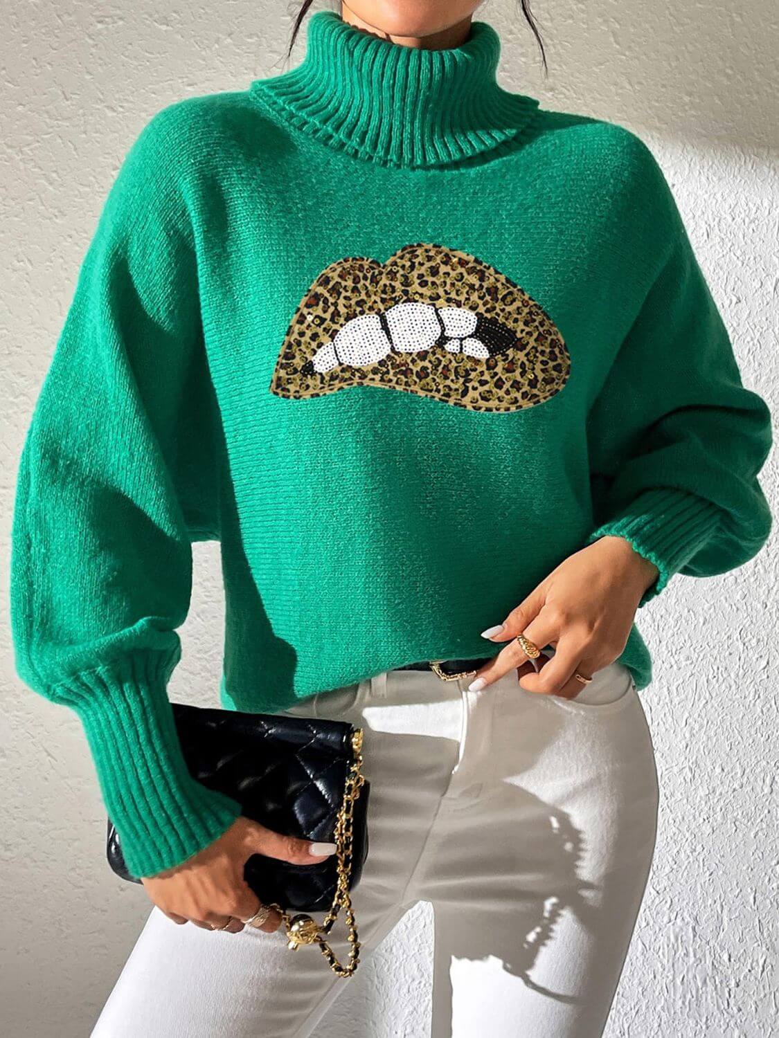 Woman wearing green Bella Road Lip Turtleneck Sweater with leopard print lips, styled with white pants and black quilted purse.