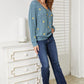 Woman wearing HEYSON floral embroidered cable cardigan with blue jeans, standing by a white dresser in sunlit room.