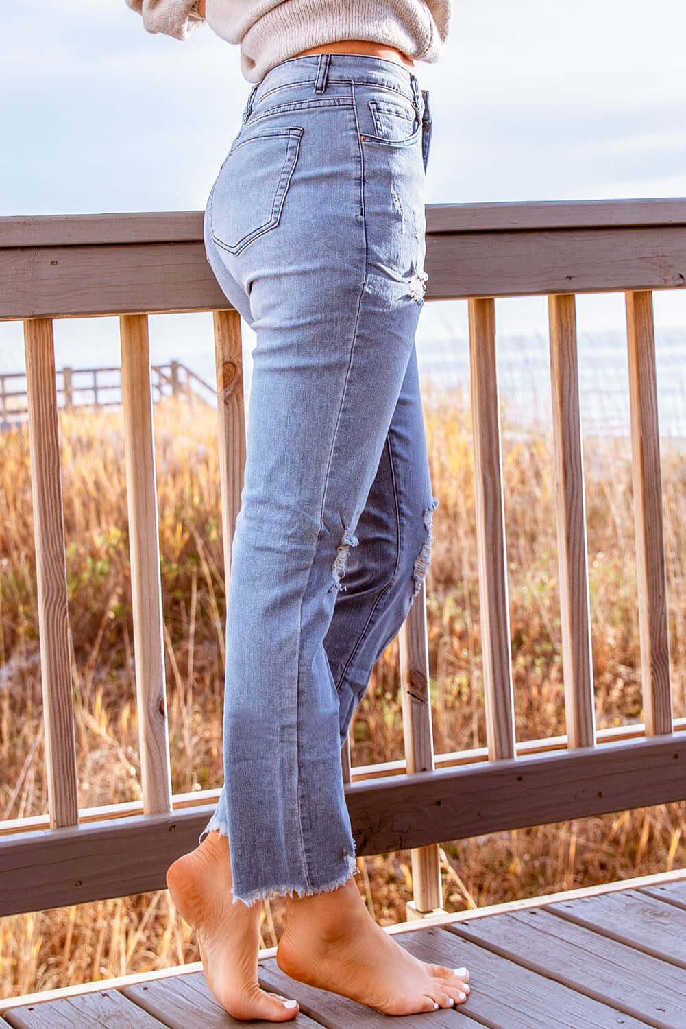 Woman wearing stylish distressed cropped jeans standing barefoot on a wooden deck by the beach.