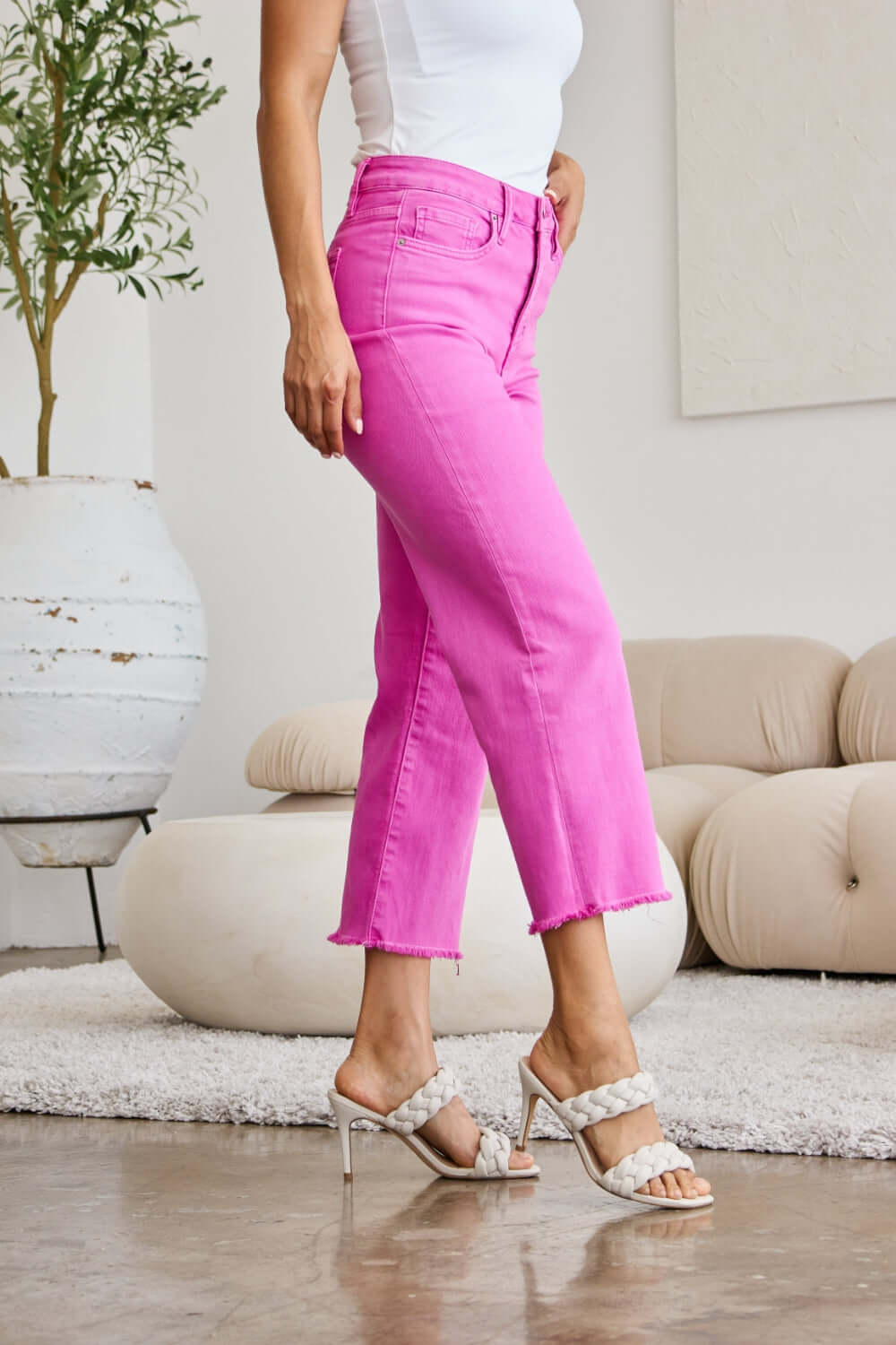 Person wearing pink RFM jeans with raw hem and high waist, styled with white tank top and heeled sandals, standing in a modern indoor setting.