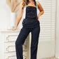 Woman wearing High Waist Classic Denim Overalls by Judy Blue Jeans standing next to a white dresser.