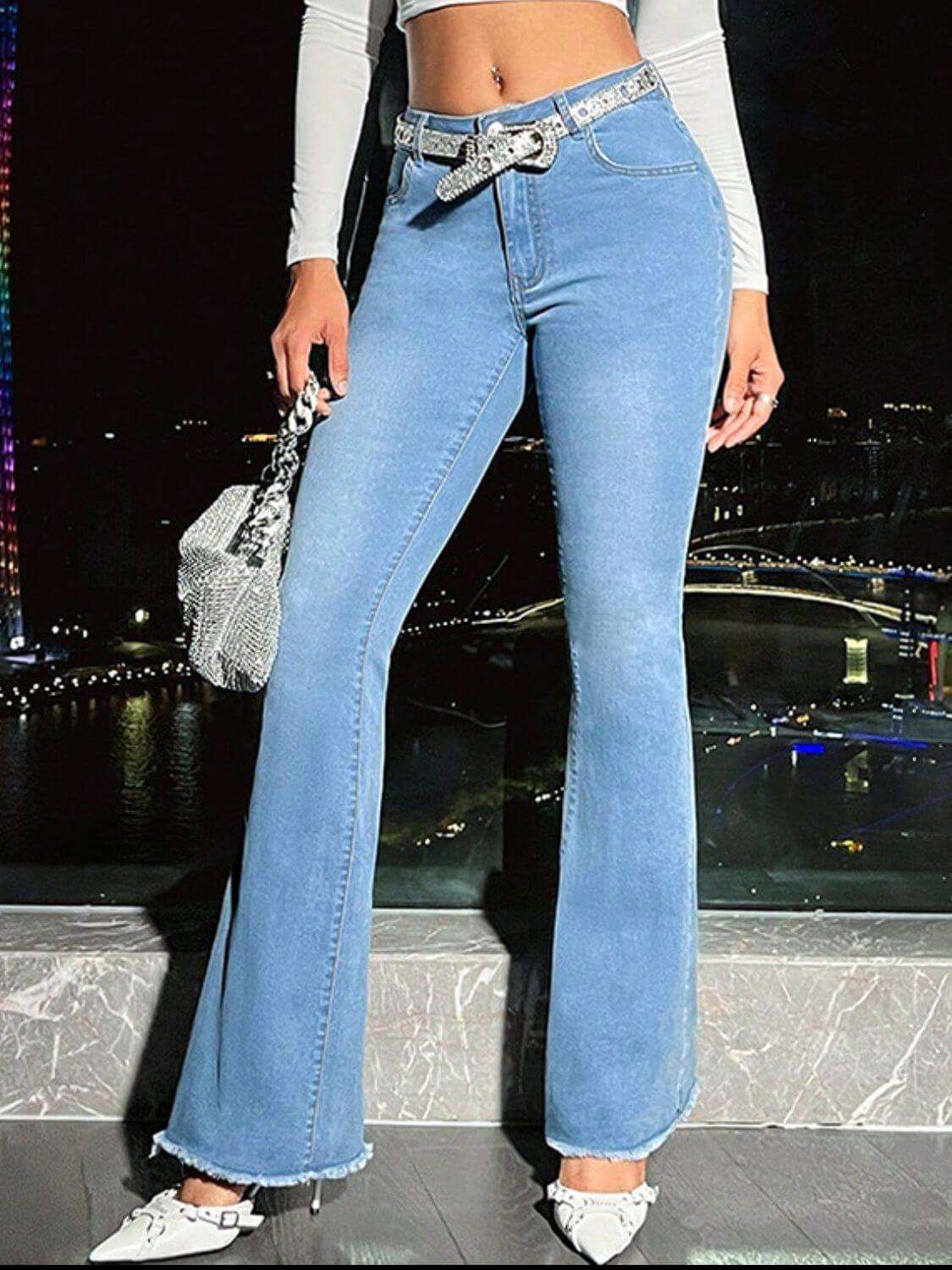 Stylish woman in light wash bootcut jeans with raw hem, accessorized with a sparkling belt and handbag.