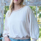 Woman wearing a light cream long-sleeve top with loose fit, standing in front of a rustic wooden backdrop.