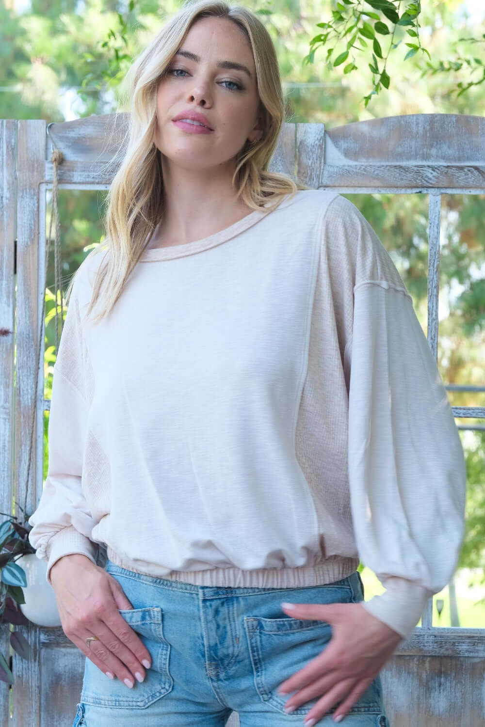 Woman wearing a light cream long-sleeve top with loose fit, standing in front of a rustic wooden backdrop.