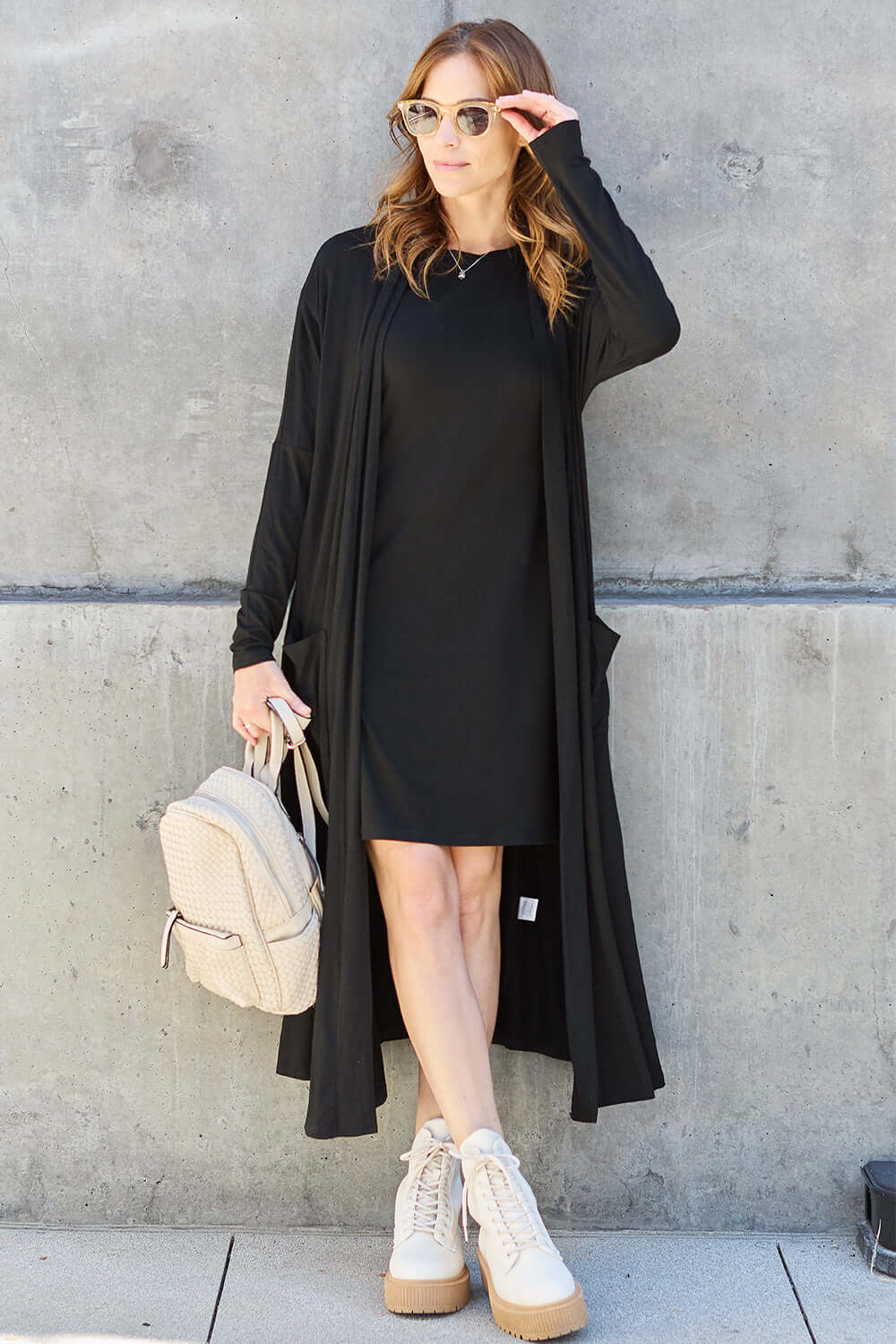 Woman wearing black open front long sleeve cover up with pockets, styled with black dress, white sneakers, sunglasses, and beige backpack.