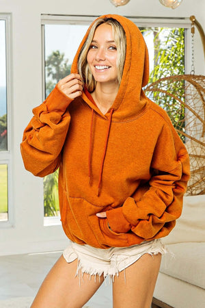 Stylish model wearing an orange BiBi Ruched Long Sleeve Washed Fleece Hoodie with casual shorts in a bright indoor setting.