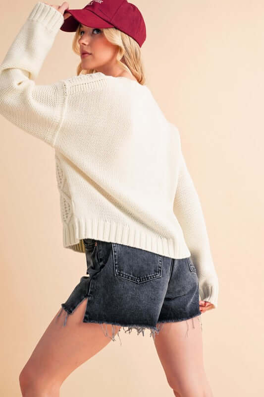 Stylish woman wearing a cozy white cable knit sweater and denim shorts, showcasing a relaxed drop shoulder silhouette.