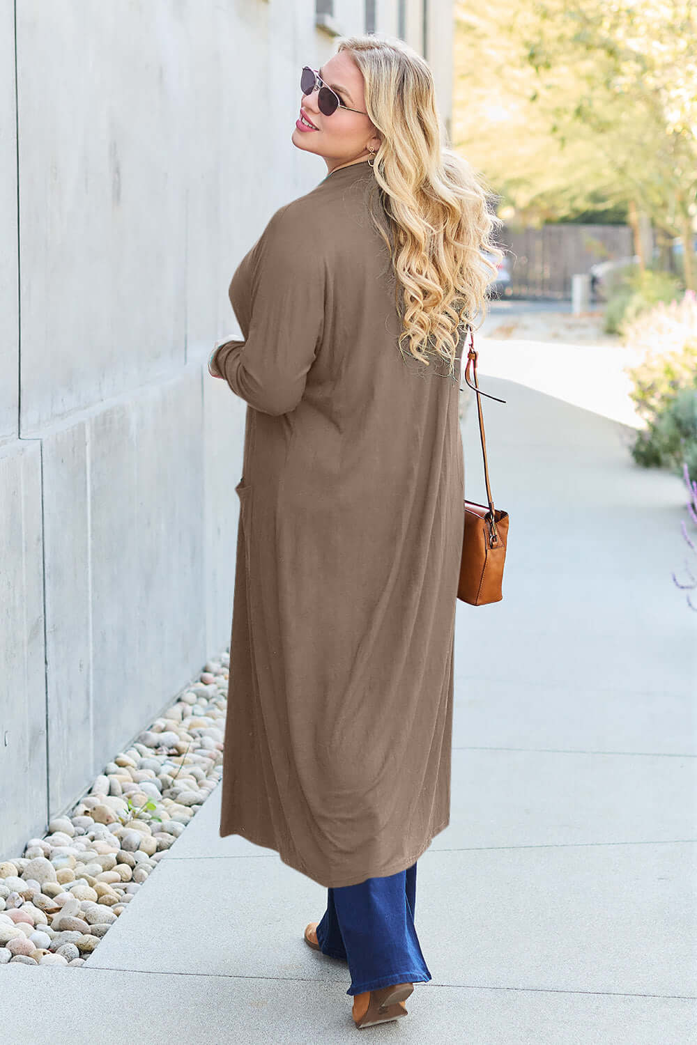 Woman wearing a brown open front long sleeve cover up with a pocket, styled with blue jeans and sunglasses, walking on a sidewalk.