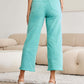 Woman wearing teal high waist raw hem jeans standing in a modern living room, Tummy Control RFM Jeans, back view for a slim and sleek silhouette