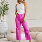 Woman wearing RFM Jeans Chloe Full Size Tummy Control High Waist Raw Hem Jeans in pink, paired with a white tank top and heels in a modern living room.