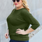 Curve model wearing a green round neck batwing sleeve top with slightly stretchy fabric and dark jeans, posing outdoors.