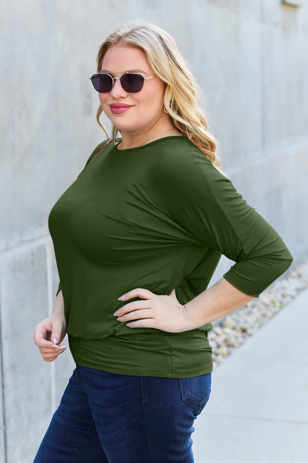 Curve model wearing a green round neck batwing sleeve top with slightly stretchy fabric and dark jeans, posing outdoors.