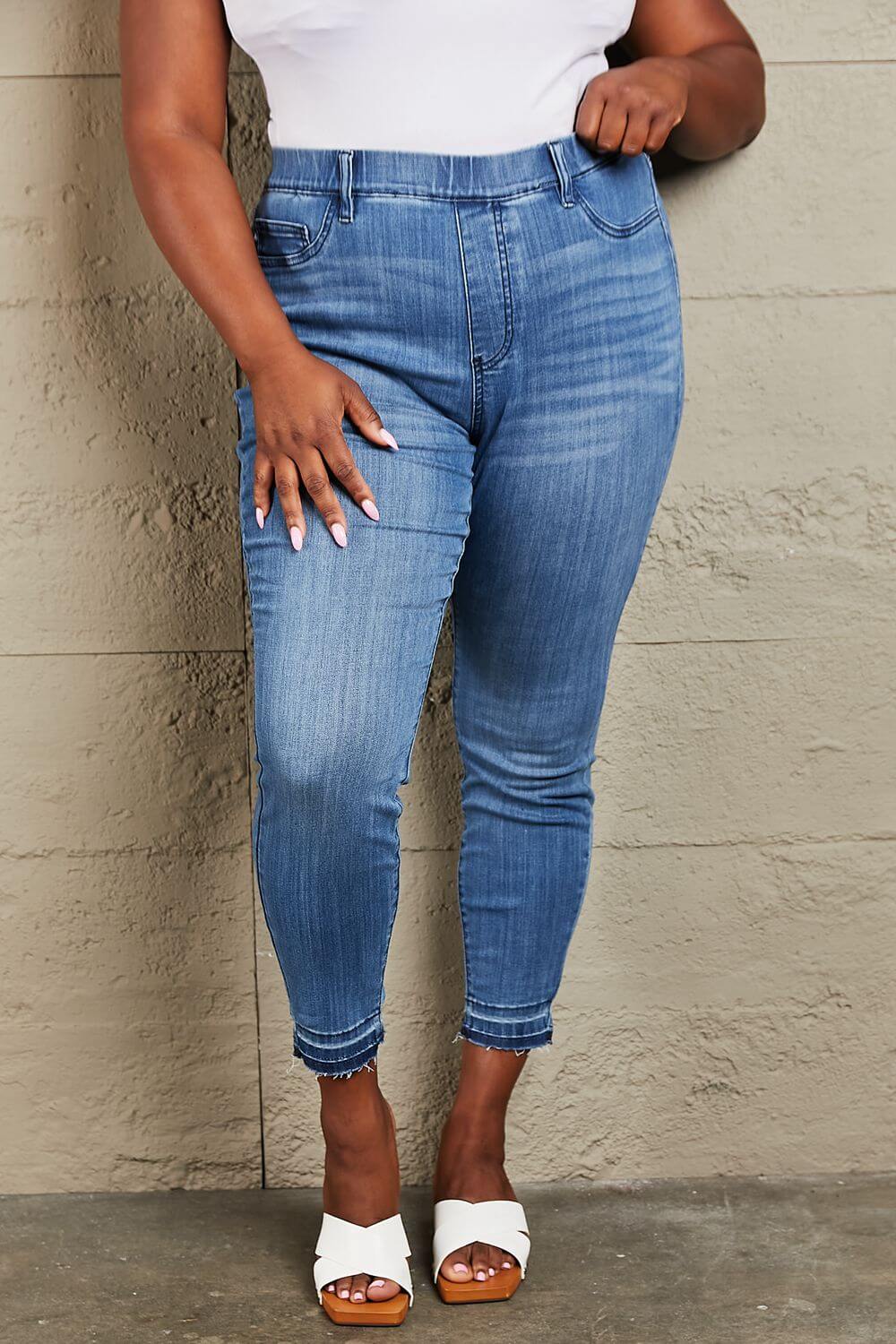 Woman wearing Janavie high waisted pull on skinny jeans with release hem detailing and white sandals. Judy Blue jeans.