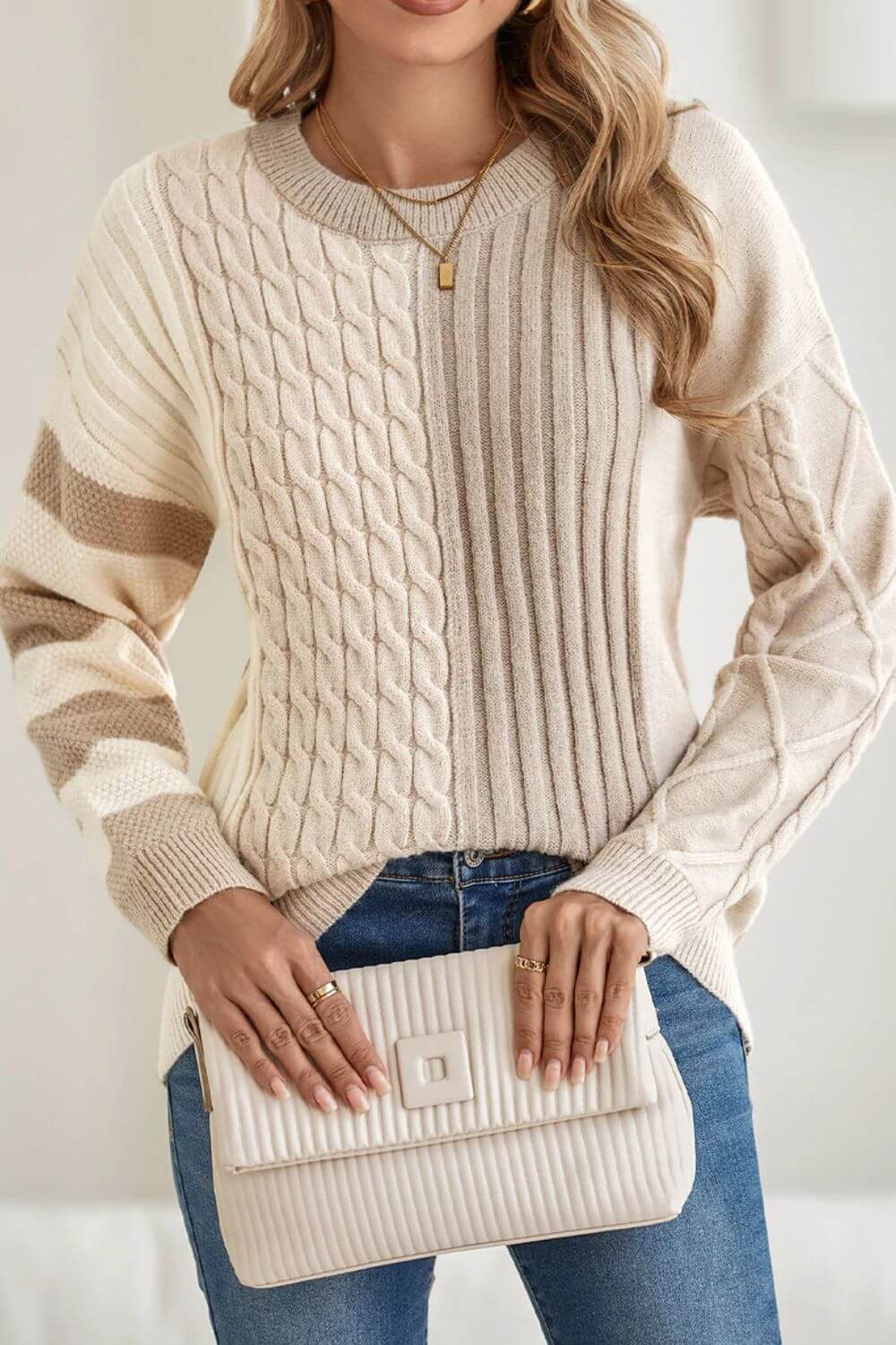 Stylish woman in Bella Road cable-knit color block round neck sweater paired with jeans, holding a chic clutch.