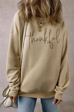 Woman wearing Bella Road Thankful Round Neck Long Sleeve Sweatshirt in beige with "thankful" text, paired with jeans and layered necklaces.