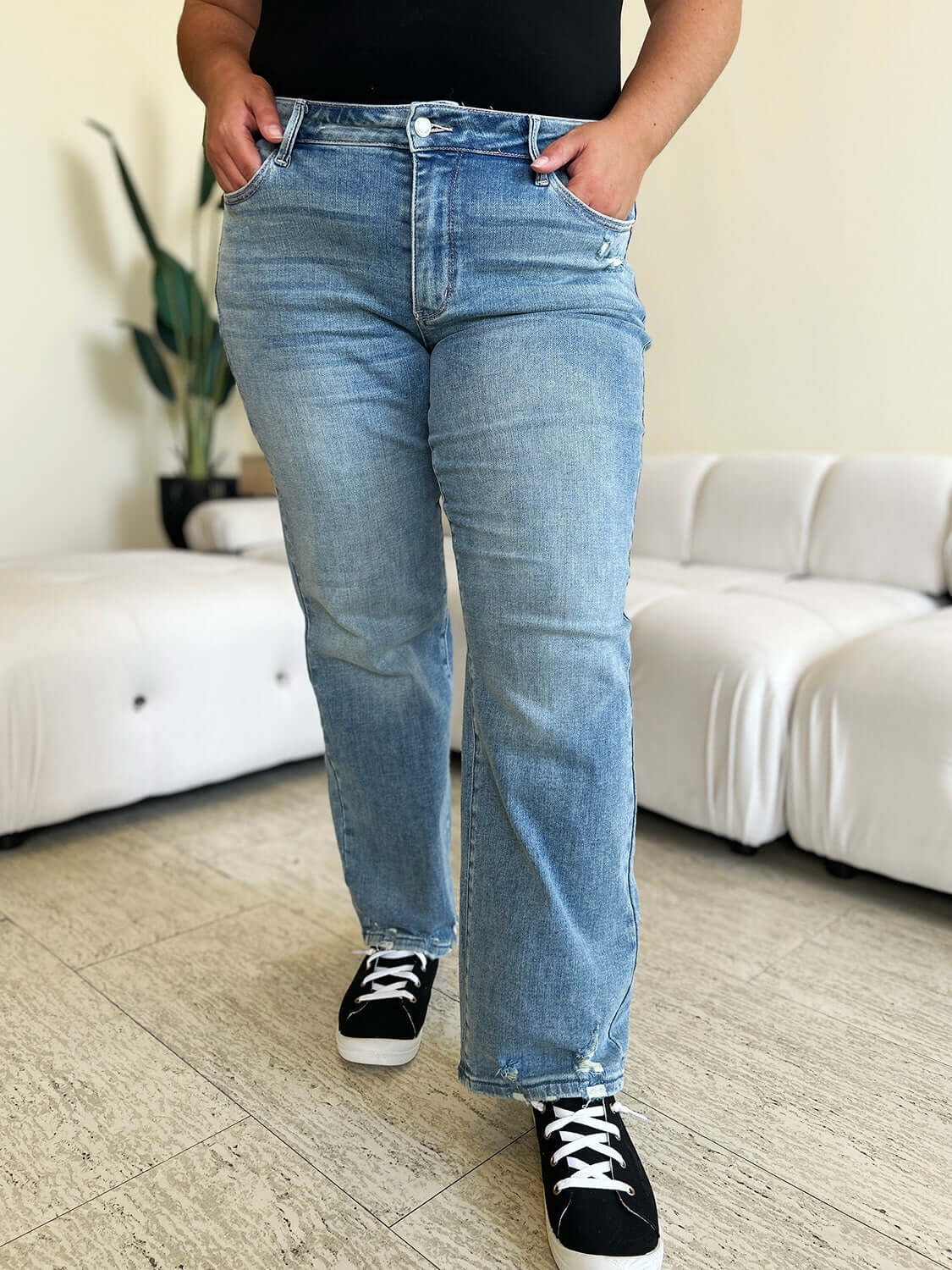 Woman wearing High Waist Distressed Straight Judy Blue Jeans with black sneakers in a modern living room.