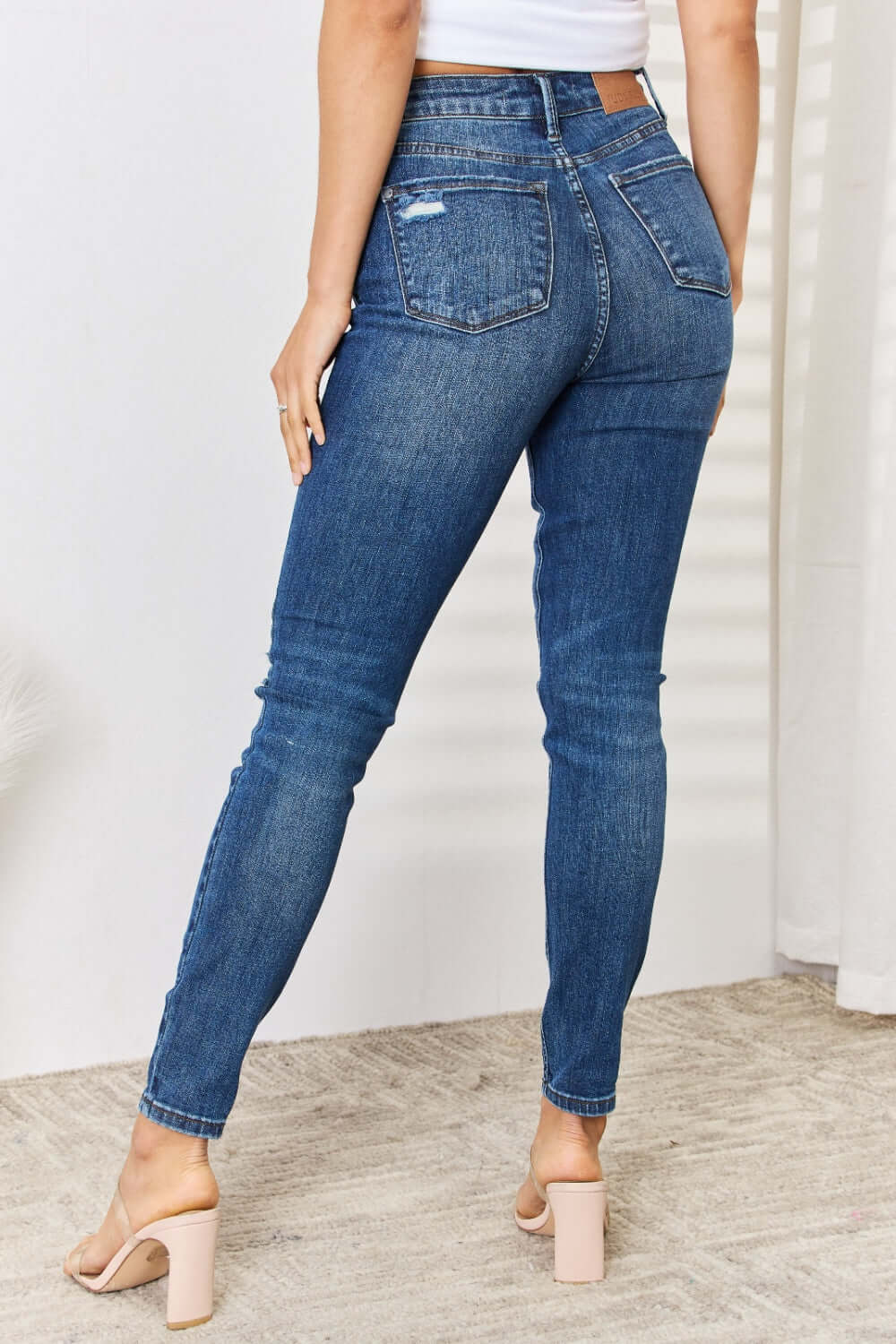 Woman wearing mid waist distressed slim jeans Judy Blue, back view showcasing flattering fit and distressed detailing, with heels