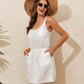 Woman wearing a white drawstring scoop neck sleeveless cover up dress, accessorized with a straw hat and sunglasses, standing against a beige wall.