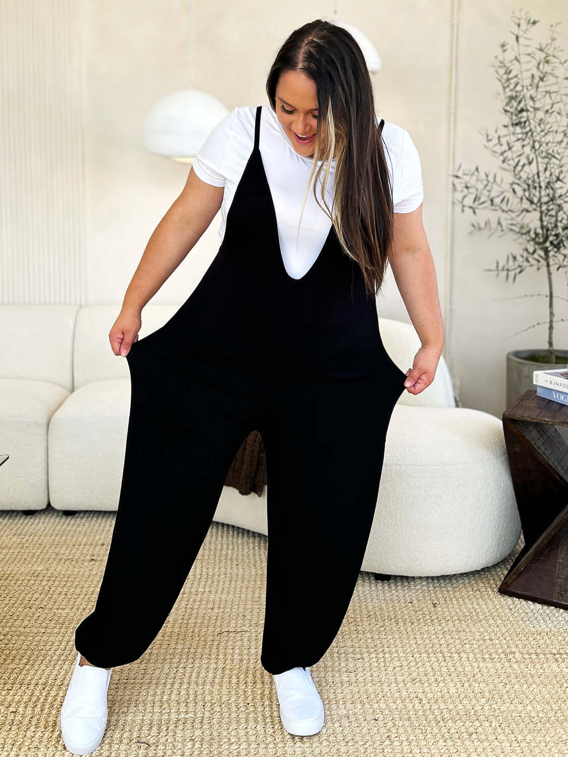 Woman wearing a black plunge sleeveless jumpsuit with pockets over white shirt in stylish living room.