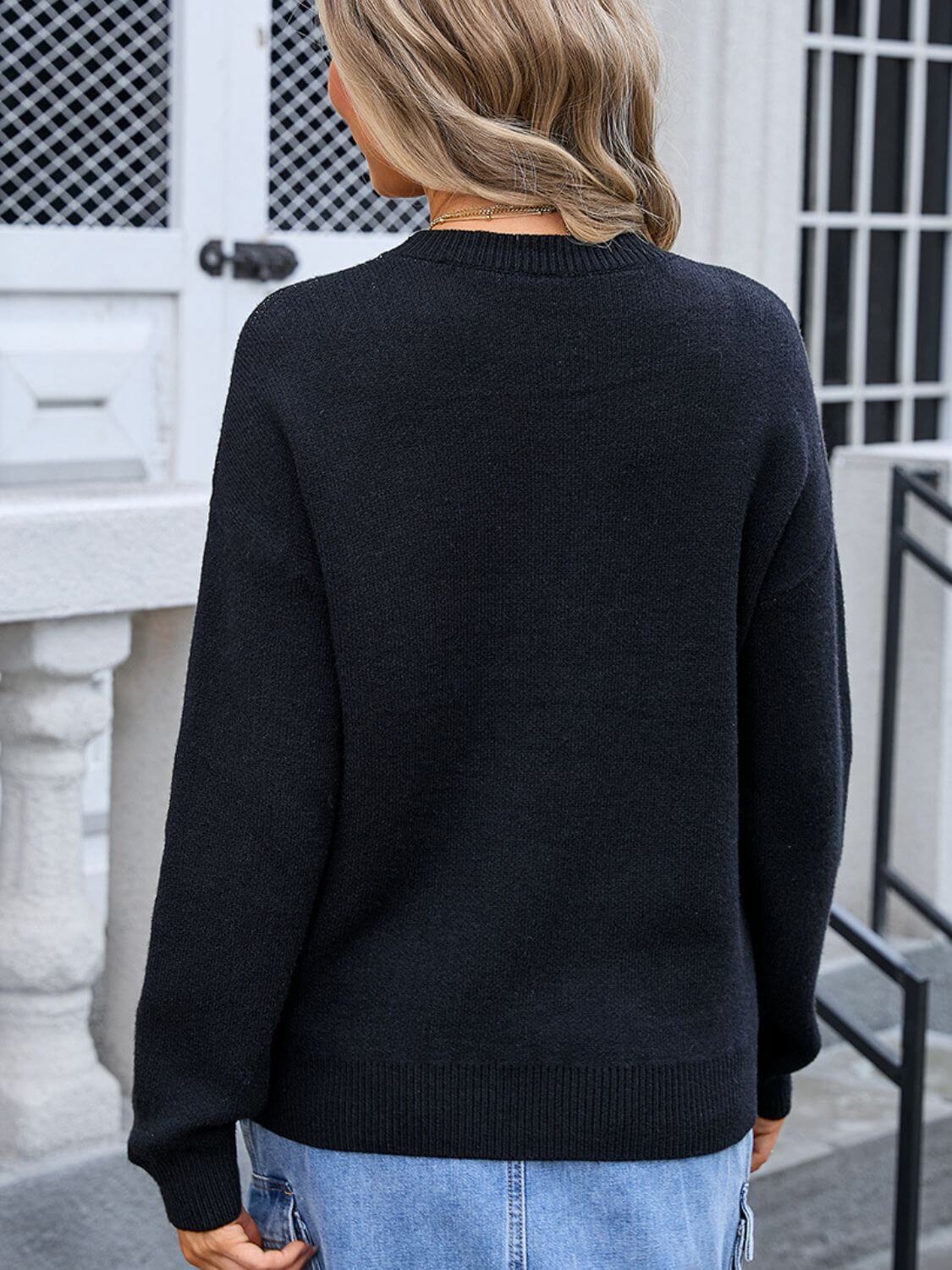 Woman wearing black round neck long sleeve sweater with angel wings bow graphic on the back, paired with denim jeans.