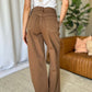 Woman wearing RFM Full Size High Rise Garment Dye Wide Leg Jeans in brown, standing indoors next to a brown sofa with green plants in the background.