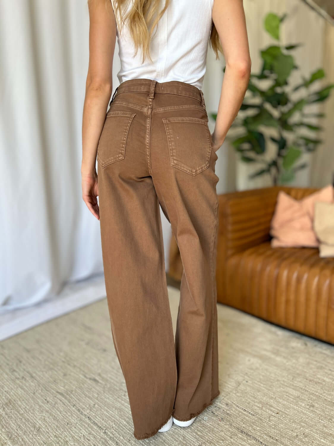 Woman wearing RFM Full Size High Rise Garment Dye Wide Leg Jeans in brown, standing indoors next to a brown sofa with green plants in the background.